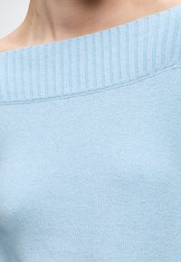 Gros plan d'une personne portant un pull côtelé bleu clair à épaules dénudées, montrant l'encolure et le haut de la poitrine.