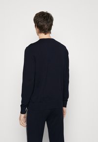 Homme de dos portant un pull noir ajusté à manches longues et un pantalon noir assorti avec des bandes blanches sur les côtés, sur un fond uni.