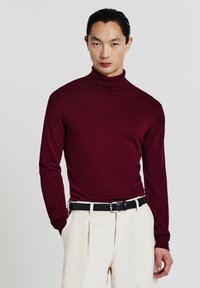 Bordeauxroter Rollkragenpullover aus weichem Strickstoff, mit langen Ärmeln, einer figurbetonten Silhouette und kombiniert mit cremefarbenen Hosen und einem schwarzen Gürtel.