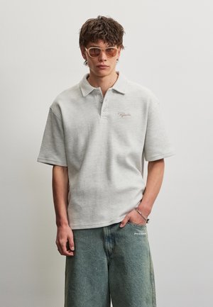 Jeune homme portant des lunettes de soleil beiges, un polo blanc texturé à manches courtes et un short vert ample, une main dans la poche, sur un fond gris uni.