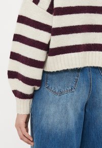 Person, der blaue Jeans und einen cremefarbenen Pullover mit dunkelvioletten horizontalen Streifen trägt, von der unteren Taille bis zur Mitte des Oberkörpers gesehen.