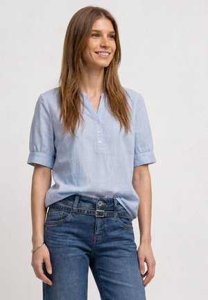 Femme souriante, portant un chemisier bleu clair à manches courtes et un jean bleu avec ceinture, debout devant un fond clair uni.