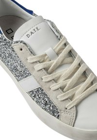 Sneaker med en overdel i hvid læder, beige ruskind detaljer og et glitrende panel i sølvglitter. Snørebåndene er tykke og cremefarvede.