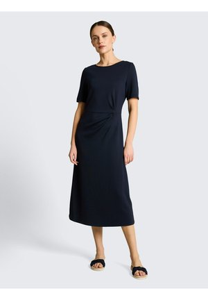 Kobieta stojąca w czarnej sukience do kolan z krótkimi rękawami i czarnych sandałach na otwarte palce, na tle jednolitego jasnego tła.