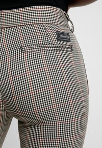 Pantalons à carreaux noir et blanc avec une poche arrière, des accents rouges et une étiquette de marque. Le tissu a une apparence texturée avec une coupe ajustée.