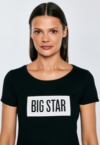 Kobieta o prostych ciemnych włosach nosi czarną koszulkę z białym prostokątnym logo z napisem "BIG STAR" na jednolitym jasnym tle.