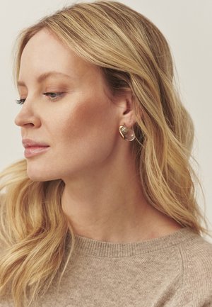 Arany karika fülbevalók átlátszó kő díszítéssel, sima, fényes felülettel. A modell világos bézs pulóvert visel.