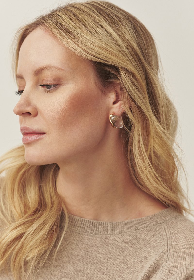 Kuldsete rõngakujuliste kõrvarõngastega, millel on läbipaistev kivielement ja sile ning läikiv viimistlus. Mudel kannab helebežikat kampsunit.