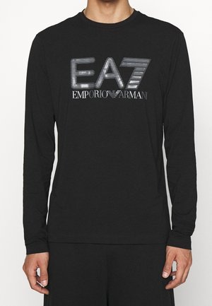 Homme portant une chemise noire à manches longues avec le logo argenté "EA7 Emporio Armani" centré sur la poitrine sur un fond uni.