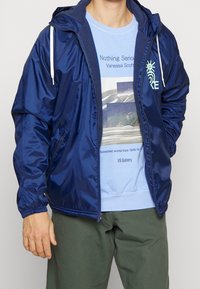 Marinblå, glänsande vindjacka med huva och vita dragsnören. Har sidofickor och elastiska ärmslut, lager-på-lager över en ljusblå sweatshirt.