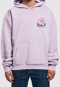 Homme portant un sweat à capuche violet clair avec poche avant et petit graphique "Sunset Charm" représentant une vague et un soleil sur le côté gauche de la poitrine, assorti à un jean bleu.