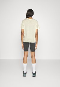 T-shirt beige chiaro a maniche corte, abbinato a pantaloni corti neri aderenti. Il modello indossa calze bianche e scarpe da ginnastica multicolori.