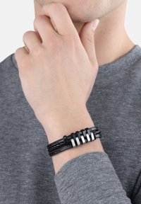 Pulsera de cuero negro con diseño trenzado y acentos de metal plateado, ajuste ajustable y superficie texturizada, llevada en una muñeca.