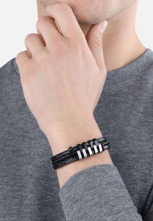 Zwarte leren armband met een gevlochten ontwerp en zilveren metalen accenten, verstelbare pasvorm en een gestructureerd oppervlak, gedragen om een pols.