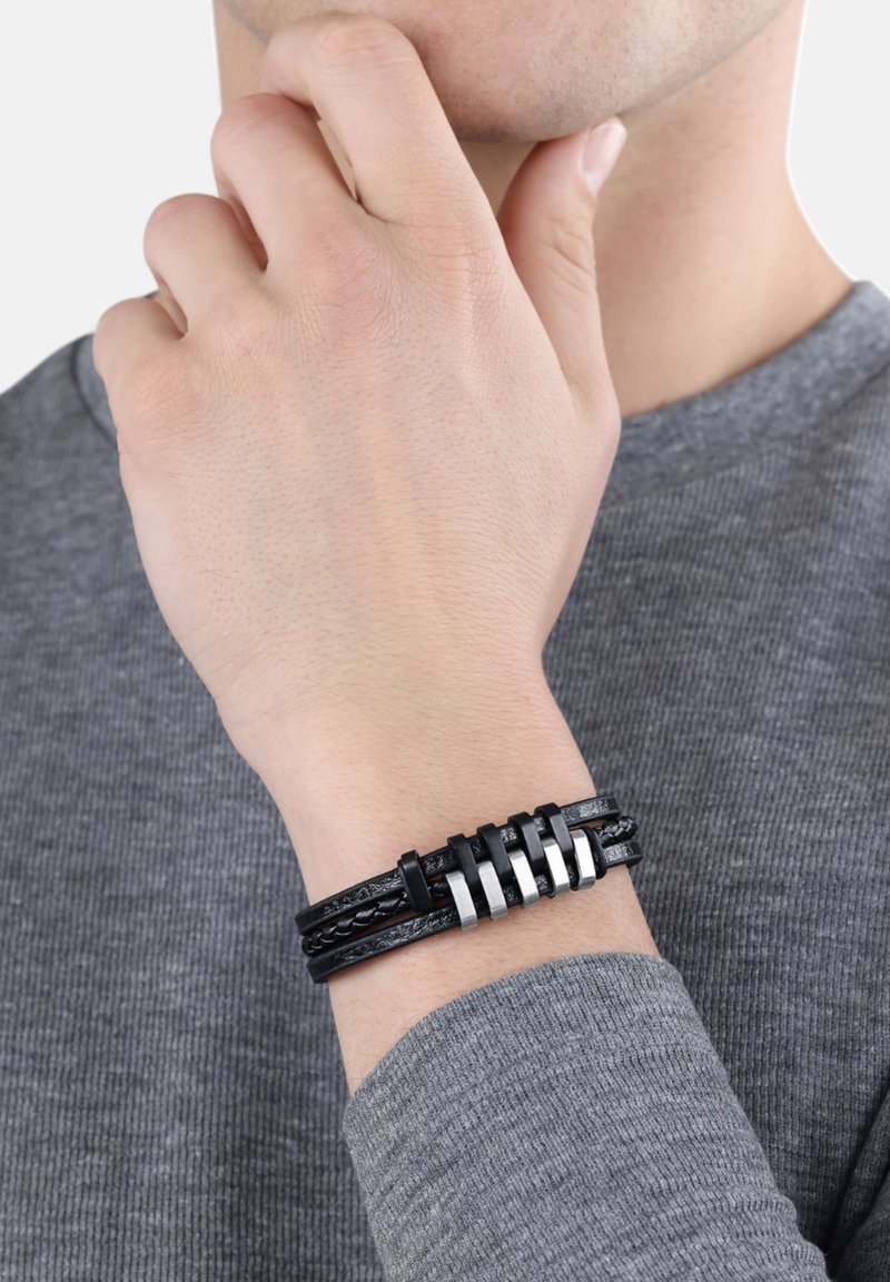 Pulsera de cuero negro con diseño trenzado y acentos de metal plateado, ajuste ajustable y superficie texturizada, llevada en una muñeca.