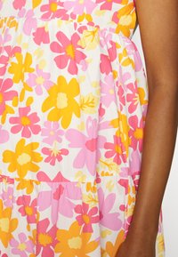 Vestido floral com grandes flores amarelas, cor-de-rosa e brancas sobre um fundo branco. Tecido texturizado com pregas na cintura.