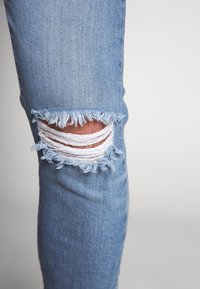 Světle modré džíny z denimu s odřeným kolenem, zdobené třepenými okraji a vystavenými bílými nitěmi. Slim fit s hladkou texturou.