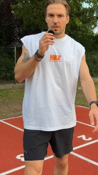 Mężczyzna w bezrękawnikowej, białej koszulce Nike i czarnych szortach stoi na zewnętrznym torze biegowym, trzymając mały czarny mikrofon blisko ust.