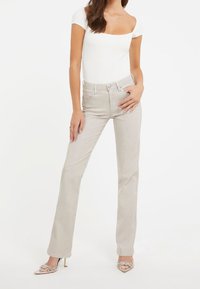 Jeans beige a vita alta con una vestibilità slim, leggermente svasati alla caviglia e design a cinque tasche; abbinati a un top aderente bianco a coste.