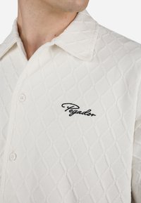 Camicia bianca a maniche corte realizzata in tessuto testurizzato, con motivo a rombi e un logo ricamato nero sul lato sinistro.