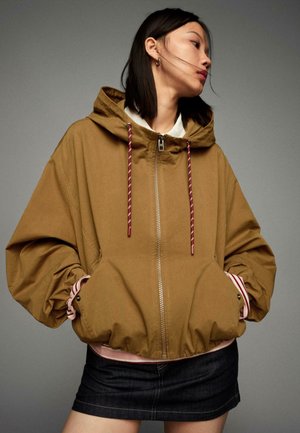 Femme portant une veste à capuche marron avec des cordons rouges, des poignets rayés et une mini-jupe en denim foncé sur fond gris uni.