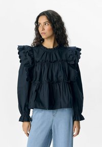 Blouse noire à volants avec des fronces superposées, des poignets élastiques et un col rond ; associée à un jean en denim bleu clair.