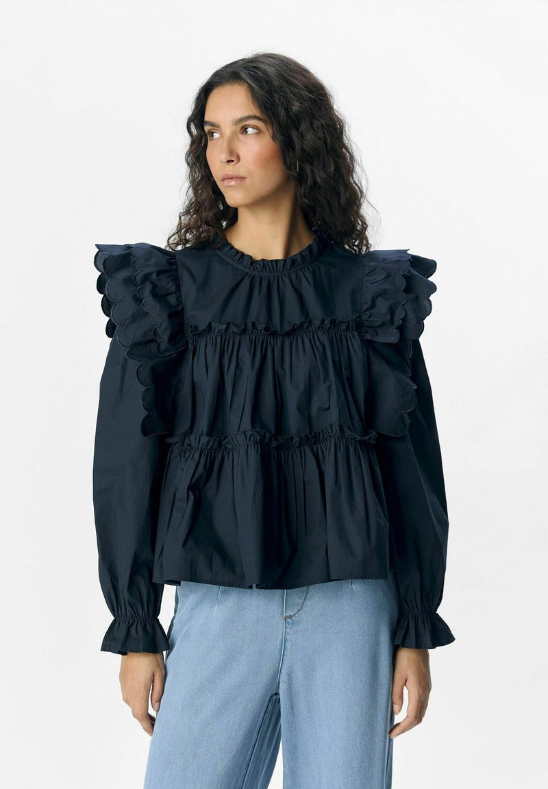 Blouse noire à volants avec des fronces superposées, des poignets élastiques et un col rond ; associée à un jean en denim bleu clair.