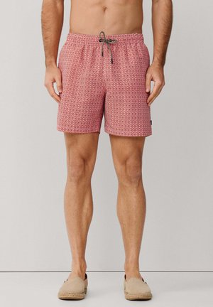 Hombre con bañador rojo con estampado y cordón negro y blanco, combinado con zapatos sin cordones beige, de pie sobre un fondo liso.