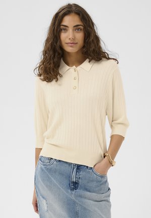 Noor naine lokkis juustega, kellel on seljas kreemjas soonikkoes polo-kampsun ja helesinine teksaseelik, käsi taskus.