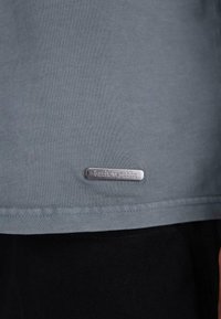 Grå t-shirt lavet af bomuld, med korte ærmer og en sølvmetal tag med indgraveret tekst nær sømmen. Glat tekstur, enkelt design.