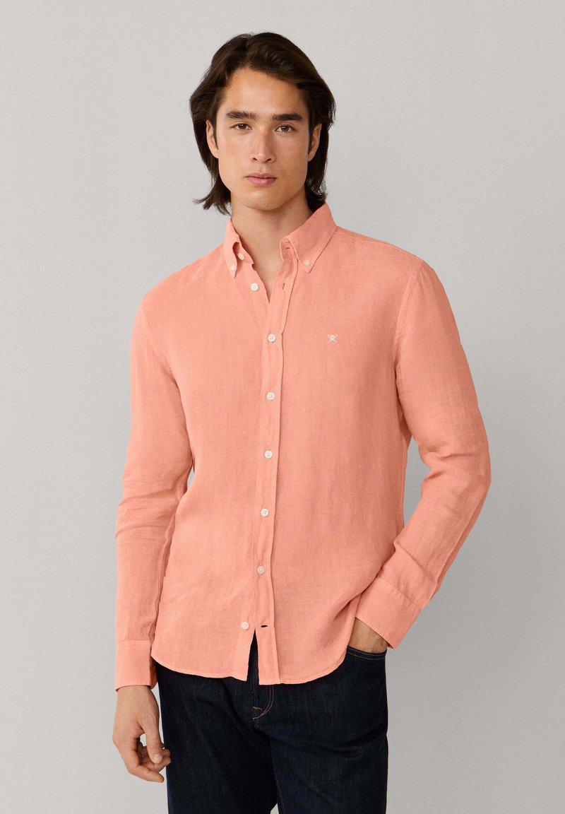 Giovane uomo che indossa una camicia a maniche lunghe color salmone con colletto e bottoni, abbinata a jeans scuri, posizionato davanti a uno sfondo neutro.