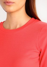 Koraliowy, prążkowany top z okrągłym dekoltem, charakteryzujący się gładką teksturą i dopasowanym krojem, podkreślający miękki, elastyczny materiał.