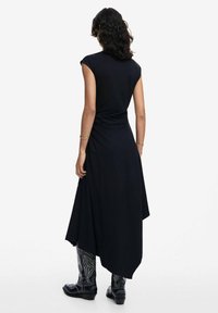 Schwarzes ärmelloses Kleid mit gerafftem Rücken, fließendem asymmetrischem Saum, das glattes Material zur Geltung bringt und mit bestickten schwarzen Stiefeln getragen wird.