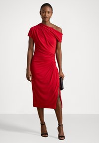 Rochie roșie, cu un umăr, cu un design plisat, până la genunchi, dintr-un material moale, cu o fanta pe lateral, purtată cu sandale negre cu toc și o geantă plic.