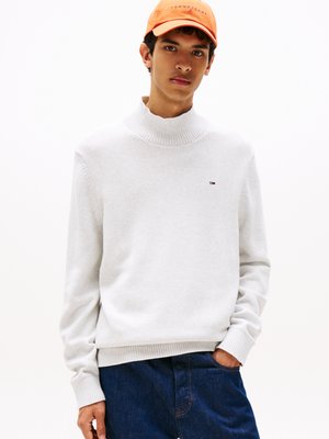 Pull gris clair avec un col côtelé haut, des poignets côtelés et un petit logo. Assorti à un jean en denim bleu foncé, créant un schéma de couleurs contrastées.
