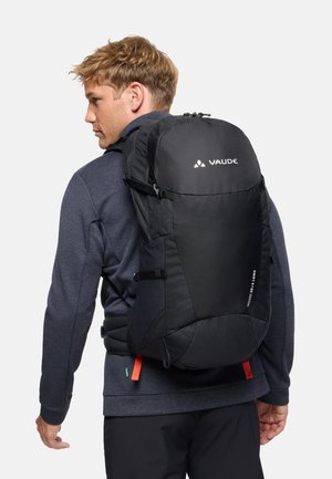 Man draagt een donker jack en broek en heeft een grote zwarte Vaude-rugzak met het label Wizard 28+4 Long op zijn rug.