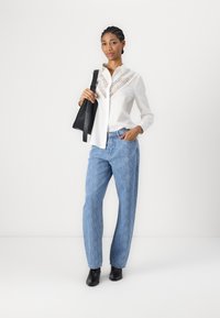 Camicia bianca con dettagli in pizzo, abbinata a jeans leggeri blu a fantasia. Il modello tiene una borsa nera e indossa stivaletti neri.