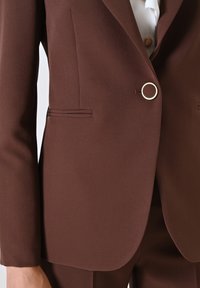 Bruine blazer van een gestructureerde stof, met een enkele ronde gouden knop, zijzakken en een getailleerde pasvorm met scherpe revers.
