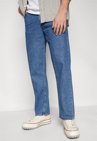 Blue denim jeans with a straight-leg fit, medium wash, and subtle texture. Paired with beige sneakers featuring red accents.