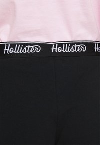 Černé legíny se širokým pasem a bílým logem "Hollister". Horní část je růžová a má hladkou texturu.