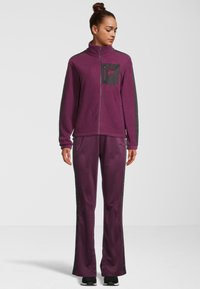 Chaqueta de forro polar morada con cuello alto, bolsillo con cremallera y detalles en negro; pantalones a juego con tiras laterales. Textura suave y corte relajado.