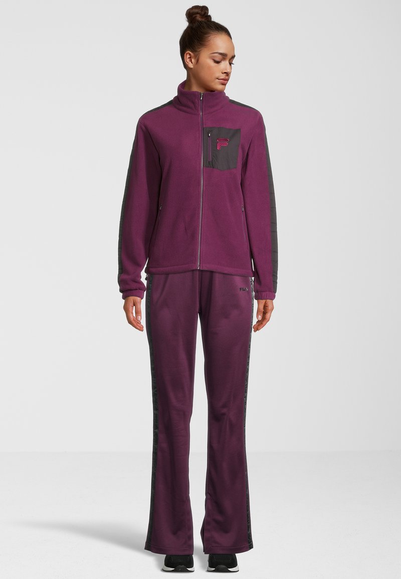 Giacca in pile viola con colletto alto, tasca con zip e dettagli neri; pantaloni coordinati con strisce laterali. Tessuto liscio e vestibilità rilassata.