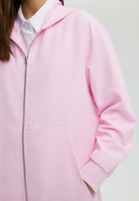 Rožā zipa hoodie, izgatavota no gludas auduma, ar kapuci, ribotām piedurknēm un priekšējo kabatu. Vienkāršs dizains, bez redzamām rakstām vai akcentiem.