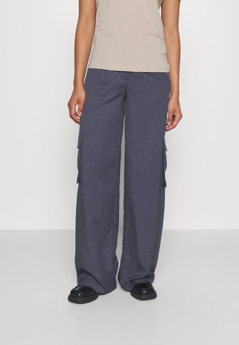 Personne portant un pantalon cargo ample gris foncé, des chaussures noires et une chemise beige à manches courtes, debout devant un fond uni.