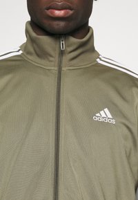 Olivovo zelená Adidas športová bunda s vysokým golierom, plným zipsom a bielymi pruhmi na pleciach. Vyznačuje sa malým bielym logom na hrudi.