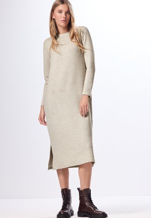 Abito lungo in maglia di un beige chiaro con collo a giro e maniche lunghe. Presenta delle aperture laterali e una silhouette rilassata. Abbinato a stivali neri con lacci.