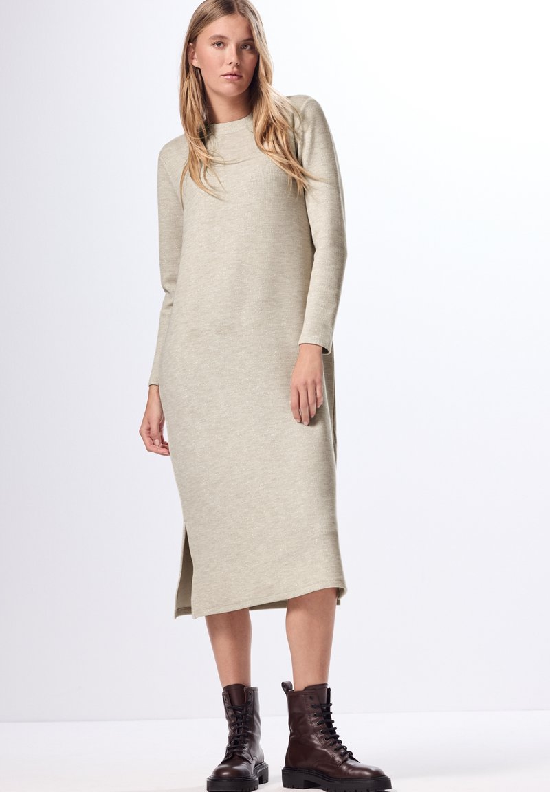 Langes, gestricktes Kleid in hellem Beige mit Rundhalsausschnitt und langen Ärmeln. Verfügt über seitliche Schlitze und eine lockere Silhouette. Kombiniert mit schwarzen Schnürstiefeln.