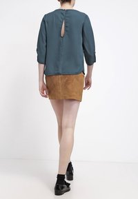 Blouse teal avec un détail en trous de serrure au dos, associée à une mini-jupe en daim marron. L'ensemble présente des manches retroussées et une texture lisse.