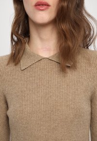Femme aux cheveux auburn ondulés, portant un pull côtelé beige avec un col pointu, lèvres légèrement entrouvertes, montrant une peau claire et des détails du cou.