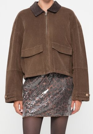 Brązowa kurtka sztruksowa na zamek błyskawiczny z dużymi przednimi kieszeniami, zestawiona z błyszczącą, wzorzystą mini spódnicą z cekinów oraz ciemnymi rajstopami.